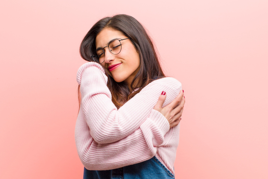 feeling in love, smiling, cuddling and hugging self, staying single, being selfish and egocentric young pretty woman feeling in love, smiling, cuddling and hugging self, staying single, being selfish and egocentric against pink background.